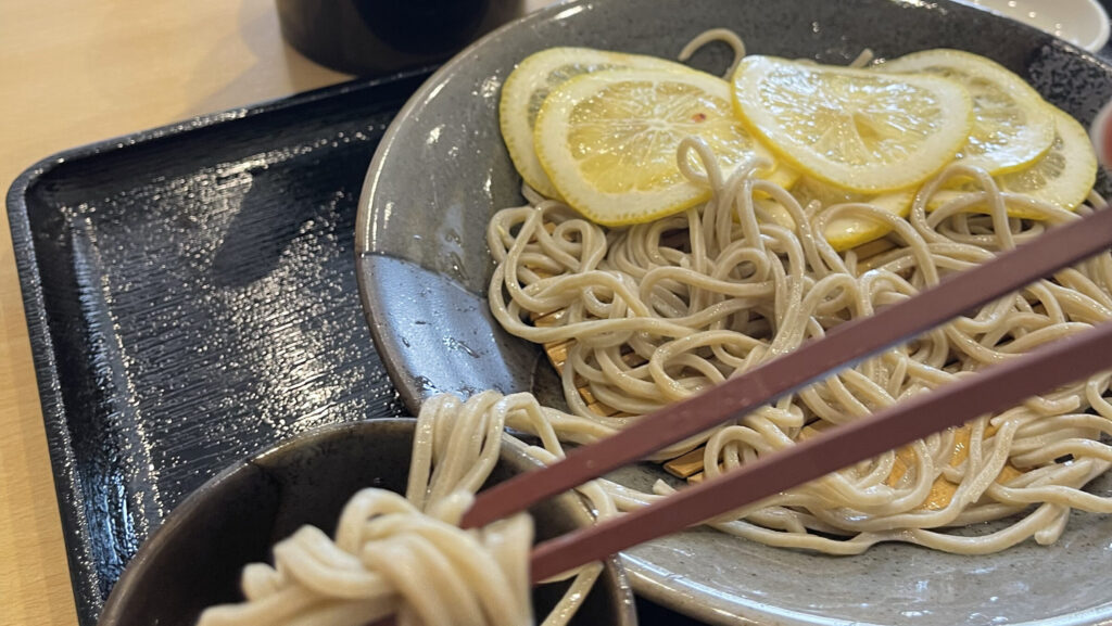 A bowl of cold soba noodles, with sliced lemon on top. Chopsticks dipping some of the noodles into a soy sauce cup are in the foreground