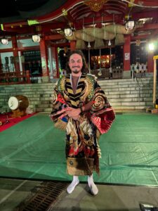 Will is wearing gold, navy and rose coloured Japanese celebration robes with a temple in the background
